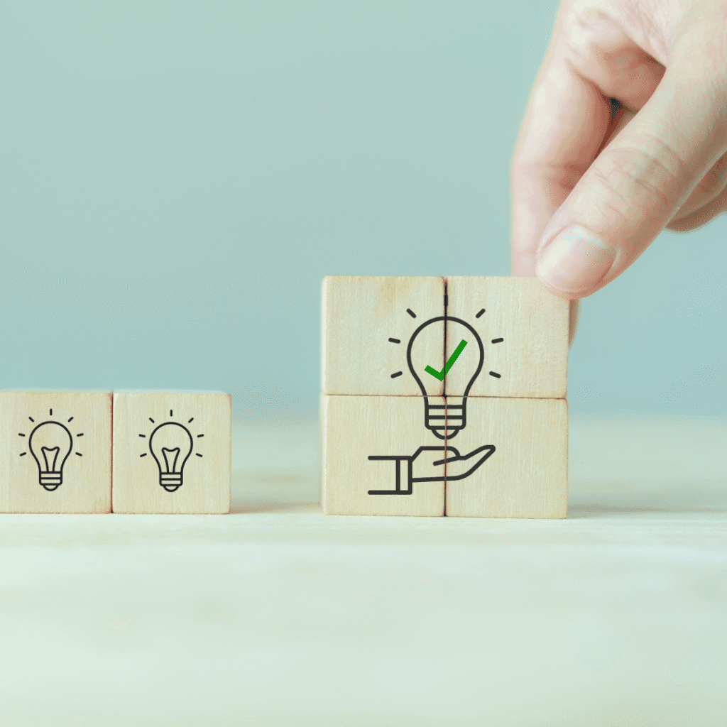 wooden blocks with light bulbs on leading to four wooden blocks being stacked together showing a lightbulb with a tick in it hovering above a hand.